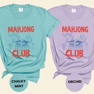 K&ouml;nnte beinhalten: Zwei T-Shirts, eines in Mintgr&uuml;n und eines in Lavendel, mit den Worten "Mahjong Club" in Rot. Die Shirts zeigen eine Grafik von Katzen, die Mahjong spielen. Das mintfarbene Shirt ist mit "Chalky Mint" und das lavendelfarbene Shirt mit "Orchid" beschriftet.