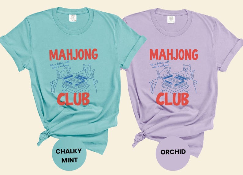 K&ouml;nnte beinhalten: Zwei T-Shirts, eines in Mintgr&uuml;n und eines in Lavendel, mit den Worten "Mahjong Club" in Rot. Die Shirts zeigen eine Grafik von Katzen, die Mahjong spielen. Das mintfarbene Shirt ist mit "Chalky Mint" und das lavendelfarbene Shirt mit "Orchid" beschriftet.