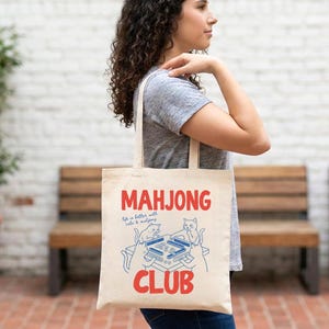 K&ouml;nnte beinhalten: Cremefarbene Tragetasche mit den roten Worten "MAHJONG CLUB" und einer blauen Illustration eines Mahjong-Tisches mit Katzen. Der Text "Life is better with cats & mahjong" ist ebenfalls enthalten.