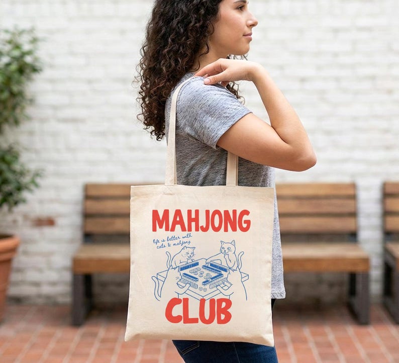 K&ouml;nnte beinhalten: Cremefarbene Tragetasche mit den roten Worten "MAHJONG CLUB" und einer blauen Illustration eines Mahjong-Tisches mit Katzen. Der Text "Life is better with cats & mahjong" ist ebenfalls enthalten.