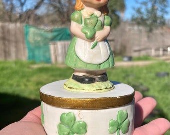 May include: A white porcelain trinket box with a green and white figurine of a girl holding shamrocks. The box is decorated with green shamrocks.