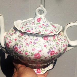 May include: A white teapot with a floral pattern in shades of pink, purple, and green. The teapot has a gold trim and a lid with a small hole in the top.
