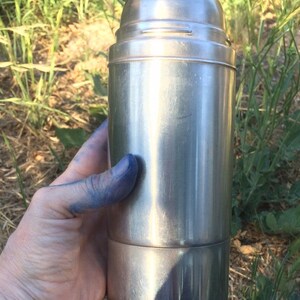 May include: A silver stainless steel thermos bottle with a screw-on lid. The bottle is being held in a hand with blue paint on the fingers.