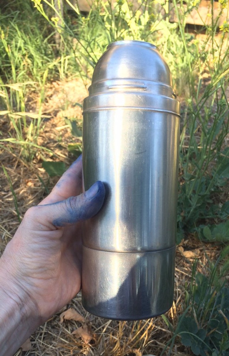 May include: A silver stainless steel thermos bottle with a screw-on lid. The bottle is being held in a hand with blue paint on the fingers.