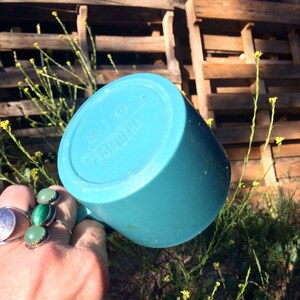 May include: A turquoise blue ceramic mug with a white inscription on the bottom. The mug is being held by a hand with green gemstone rings.