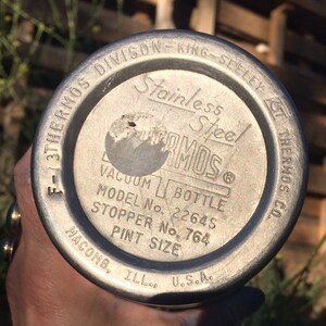 May include: Close-up of the bottom of a silver thermos with the text "3THERMOS DIVISON-KING-SEELEY F-1 Stainless Steel THERMOS VACUUM BOTTLE MODEL No. 2264S STOPPER No. 764 PINT SIZE MACOMB. ILL. U.S.A. CO."