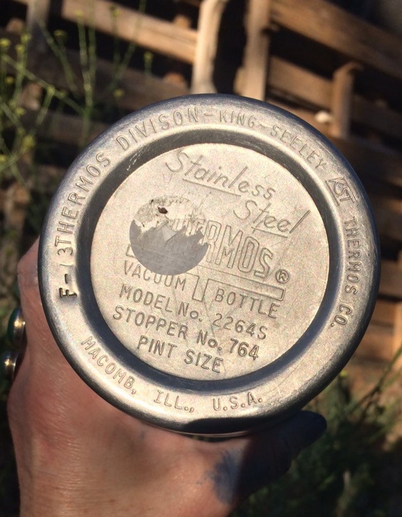 May include: Close-up of the bottom of a silver thermos with the text "3THERMOS DIVISON-KING-SEELEY F-1 Stainless Steel THERMOS VACUUM BOTTLE MODEL No. 2264S STOPPER No. 764 PINT SIZE MACOMB. ILL. U.S.A. CO."