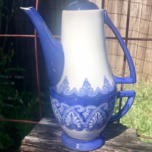 May include: A vintage ceramic coffee pot with a white body and blue accents. The pot features a curved spout, a loop handle, and a decorative blue pattern around the base. The lid is dark blue.