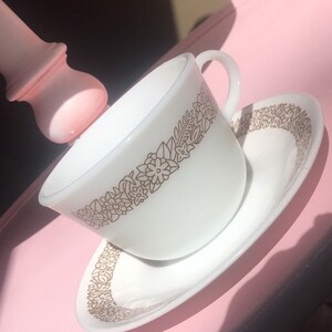 May include: A white teacup and saucer with a gold floral pattern. The cup and saucer are on a pink surface.