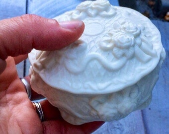 May include: White ceramic trinket box with a raised floral design. The box has a lid with a scalloped edge and a smooth, white finish.