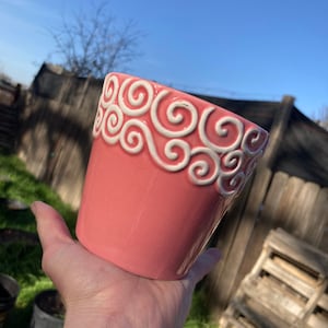 May include: A pink ceramic planter with a white swirl design around the top.