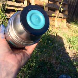 May include: A stainless steel water bottle with a turquoise blue lid and a black rubber ring. The bottle is sitting on a wooden pallet.