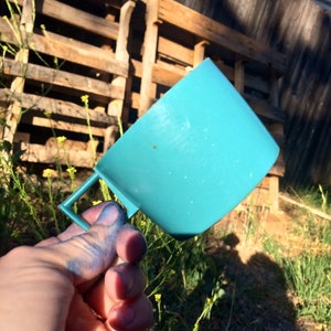 May include: A teal blue plastic measuring cup with a handle. The cup is being held by a hand with blue paint on the fingers.
