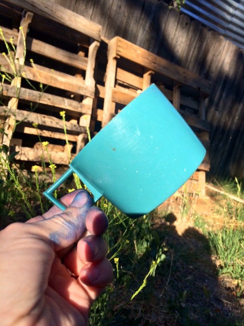 May include: A teal blue plastic measuring cup with a handle. The cup is being held by a hand with blue paint on the fingers.