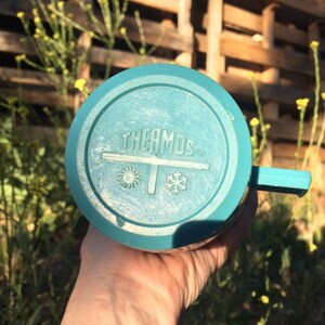 May include: A teal blue thermos with a white lid and a white label that says "THERMOS" with a snowflake and sun symbol on either side of the label.