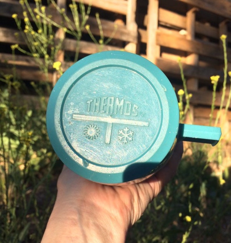 May include: A teal blue thermos with a white lid and a white label that says "THERMOS" with a snowflake and sun symbol on either side of the label.