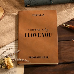 May include: A tan leather journal with the text "reasons why I LOVE YOU". The journal is personalised with "TO: VIVIAN" at the top and "FROM: MICHAEL" at the bottom. Other journals and pens are visible in the background.