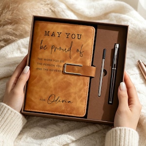 Pode incluir: Um diário em imitação de couro castanho com um fecho metálico dourado, com o texto "MAY YOU be proud of THE WORK YUO DO THE PERSON YOU A AND THE DIFFEREN" e "FOR Olivia". O diário está numa caixa com uma caneta preta e uma recarga.