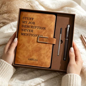 Puede incluir: Un cuaderno de piel sintética color canela con un cierre de metal dorado, que presenta el texto STUFF MY JOB DESCRIPTION NEVER MENTIONED y el nombre AMELIA. Se presenta en una caja marrón junto a un bolígrafo negro y un recambio plateado.