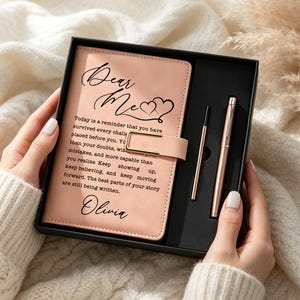 Puede incluir: Un diario de color melocotón claro con cierre magnético, que presenta el texto "Dear Me" y una cita inspiradora, personalizado con el nombre "Olivia". El set incluye un bolígrafo de oro rosa a juego y un recambio, en una caja negra.