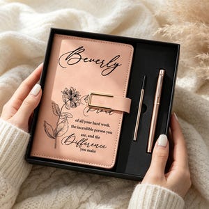 May include: A light pink notebook with a gold-tone magnetic clasp, featuring a black floral illustration and the text "Beverly, Proud of all your hard work, the incredible person you are, and the Difference you make," paired with a matching pen.