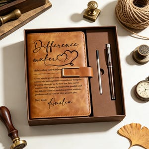 May include: A tan leather-like journal with a gold-tone metal clasp, featuring the printed text Difference maker and a definition, alongside a matching pen and a refill cartridge, all presented in a brown rectangular gift box.