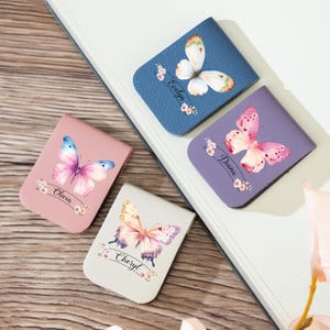 Może przedstawiać: Cztery magnetyczne zakładki do książek z motywem motyla. Każda zakładka ma inny wzór motyla i imię: Evelyn, Olivia, Diana i Cheryl. Zakładki są w odcieniach niebieskiego, różowego, fioletowego i złamanej bieli.