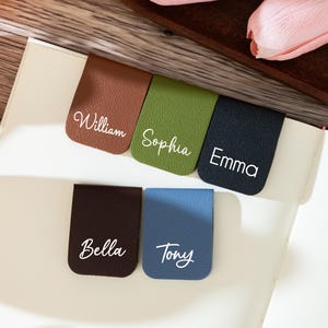 Może przedstawiać: Kolekcja spersonalizowanych skórzanych zakładek do książek w różnych kolorach, w tym brązowym, zielonym i niebieskim. Każda zakładka zawiera imię w białym skrypcie: William, Sophia, Emma, Bella i Tony. Zakładki umieszczone są na stronach otwartej książki.