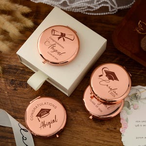 May include: Rose gold compact mirrors with personalized graduation messages. Each mirror features a unique design, including a graduation cap and scroll, along with the graduate's name and the year 2026. The mirrors are presented in a white gift box.