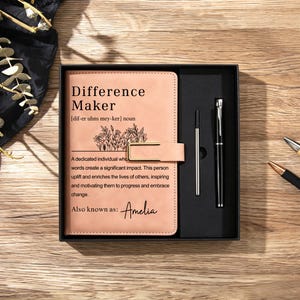 May include: A light tan leather-bound journal with a gold metal clasp, featuring the printed text Difference Maker and a floral illustration, presented in a black gift box alongside a black metal pen and a silver ink refill cartridge.
