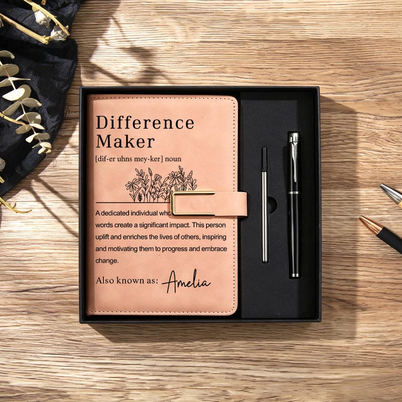 May include: A light tan leather-bound journal with a gold metal clasp, featuring the printed text Difference Maker and a floral illustration, presented in a black gift box alongside a black metal pen and a silver ink refill cartridge.