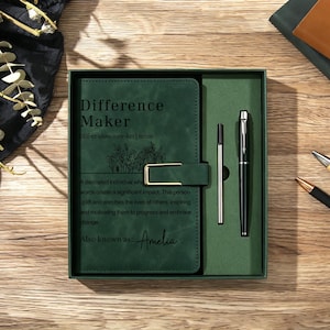 May include: A dark green notebook with a gold-toned metal clasp and a matching black ink pen in a gift box. The notebook cover features the text Difference Maker, a definition, and the name Amelia printed in black ink with a floral line drawing.