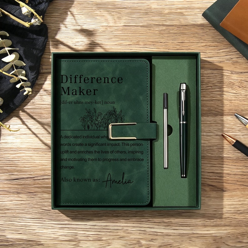 May include: A dark green notebook with a gold-toned metal clasp and a matching black ink pen in a gift box. The notebook cover features the text Difference Maker, a definition, and the name Amelia printed in black ink with a floral line drawing.