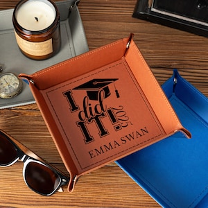 Puede incluir: Una bandeja de cuero sintético marrón con el texto "I did it! 2023 Emma Swan" y un gráfico de gorro de graduación. La bandeja es cuadrada con esquinas plegadas. Se ven otras bandejas en gris y azul.