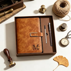 May include: A tan notebook with a gold-toned metal clasp and the name Mateo printed in black, presented in a brown gift box alongside a matching brown pen and a single ink refill. The notebook cover features a large letter M monogram design.