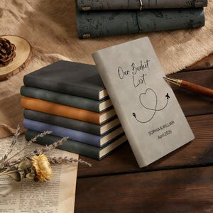 May include: A stack of journals in various colors, including gray, brown, and blue, with a light gray journal in the foreground. The light gray journal has the text "Our Bucket List" and a heart-shaped travel route design.