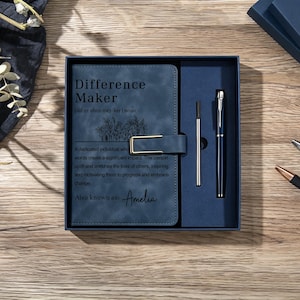 May include: A dark blue notebook with a gold-toned metal clasp and the text Difference Maker, a definition, and the name Amelia printed on the cover. The notebook is presented in a matching blue gift box alongside a metal pen and a refill cartridge.