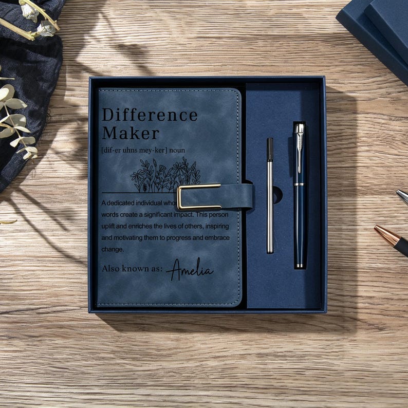 May include: A dark blue notebook with a gold-toned metal clasp and the text Difference Maker, a definition, and the name Amelia printed on the cover. The notebook is presented in a matching blue gift box alongside a metal pen and a refill cartridge.