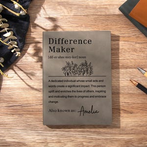 May include: A rectangular grey notebook featuring the printed text Difference Maker, a phonetic pronunciation guide, a line drawing of wildflowers, and a definition. The text also includes the phrase Also known as: Amelia in script font.
