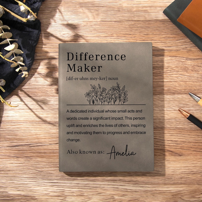 May include: A rectangular grey notebook featuring the printed text Difference Maker, a phonetic pronunciation guide, a line drawing of wildflowers, and a definition. The text also includes the phrase Also known as: Amelia in script font.