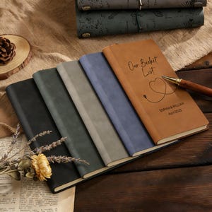May include: A collection of leather-bound journals in various colors, including black, gray, and brown. One journal is open, displaying the text "Our Bucket List" with a heart design and the names "Sophia & William" and the date "April 2025".