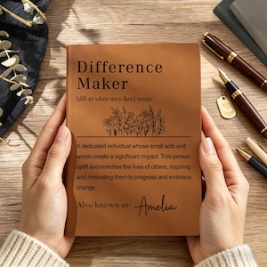 May include: A tan leather-bound notebook featuring the printed text Difference Maker, a phonetic pronunciation, a line drawing of wildflowers, and a definition of the term. The bottom of the cover includes the printed name Amelia in script font.
