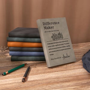 May include: A stack of seven rectangular notebooks with matte covers in grey, dark green, orange, black, blue, and dark grey. The top grey notebook features the text Difference Maker, a definition, a floral illustration, and the name Amelia.