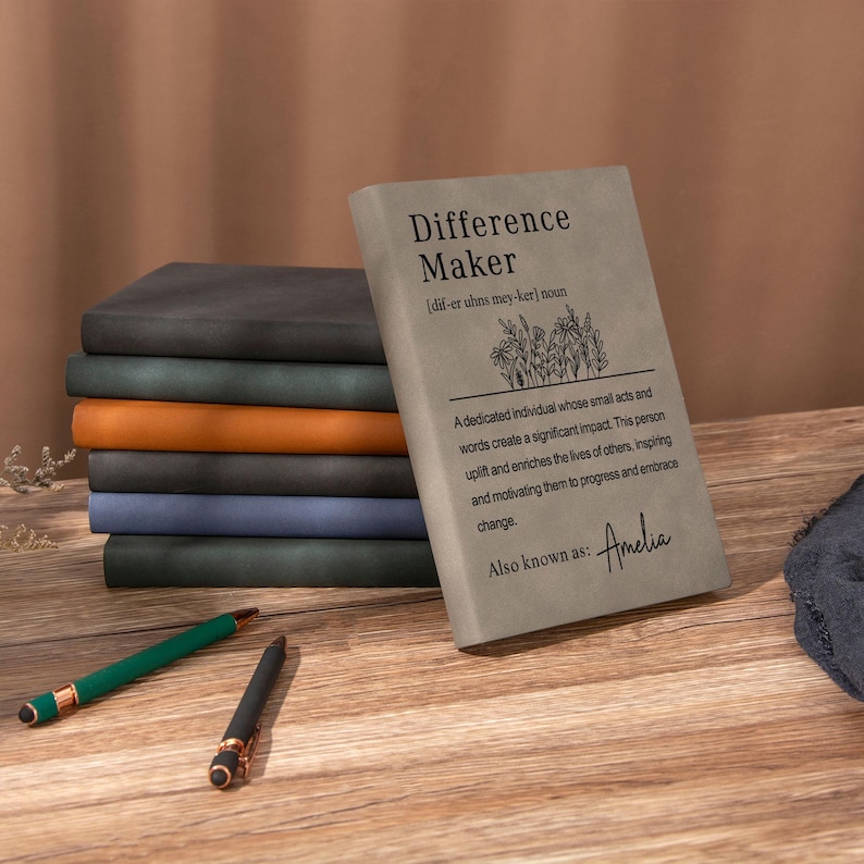 May include: A stack of seven rectangular notebooks with matte covers in grey, dark green, orange, black, blue, and dark grey. The top grey notebook features the text Difference Maker, a definition, a floral illustration, and the name Amelia.