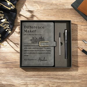 May include: A grey notebook with a gold-coloured metal clasp and a matching black pen set in a gift box. The cover features the text Difference Maker, a floral illustration, a definition, and the name Amelia printed in black ink on the front.