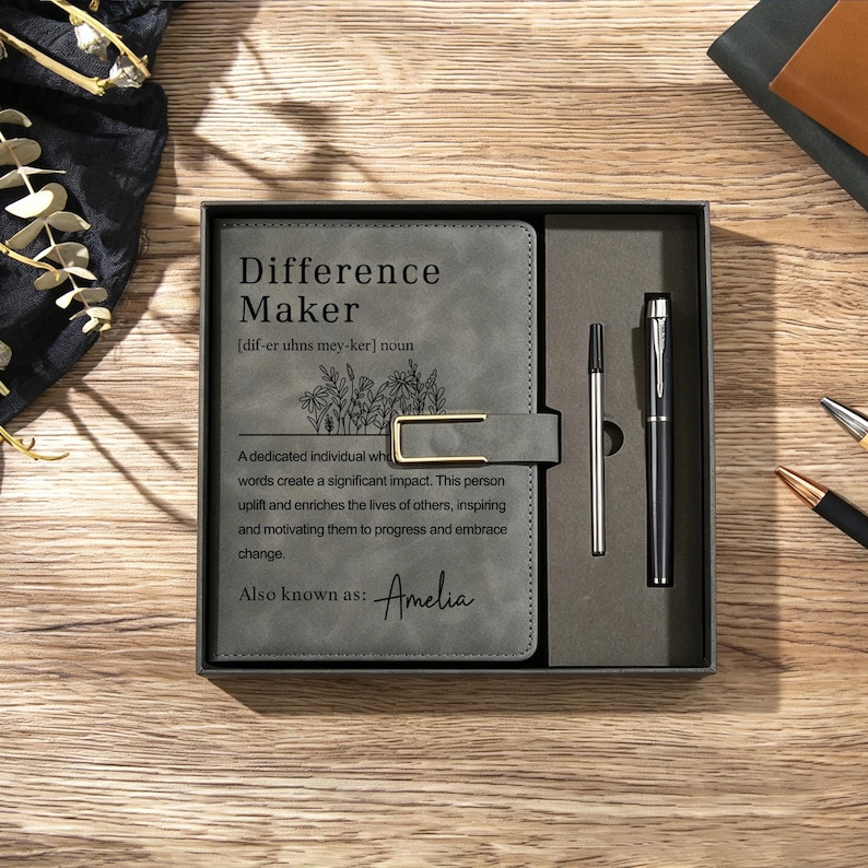 May include: A grey notebook with a gold-coloured metal clasp and a matching black pen set in a gift box. The cover features the text Difference Maker, a floral illustration, a definition, and the name Amelia printed in black ink on the front.
