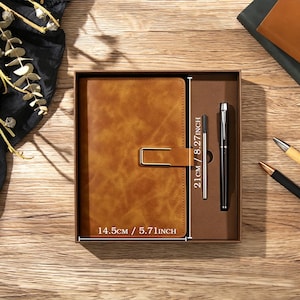 May include: A tan notebook with a gold-tone metal clasp and a black pen in a brown gift box. The notebook measures 14.5 cm by 21 cm. The set includes a pen refill. The box is presented on a wooden surface with decorative elements.