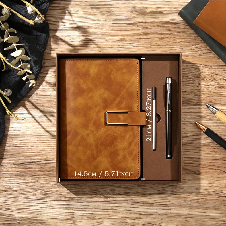 May include: A tan notebook with a gold-tone metal clasp and a black pen in a brown gift box. The notebook measures 14.5 cm by 21 cm. The set includes a pen refill. The box is presented on a wooden surface with decorative elements.