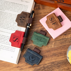 Op de afbeelding: Een verzameling gepersonaliseerde leren penhouders in verschillende kleuren, waaronder bruin, rood, groen en blauw. Elke houder heeft een naam of initialen en is bevestigd aan een pen of notitieblok. De items zijn gerangschikt op een houten oppervlak.