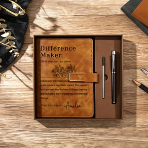 May include: A tan leather-look journal with a gold metal clasp, featuring the printed text Difference Maker, a floral line drawing, and the name Amelia. The journal is housed in a brown gift box alongside a black pen and a silver ink refill.